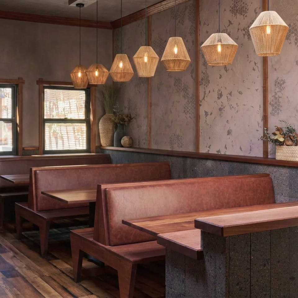 Warm restaurant dining area with wooden booths, textured walls, hanging wicker lights, decorative vases, and sunlight through windows.