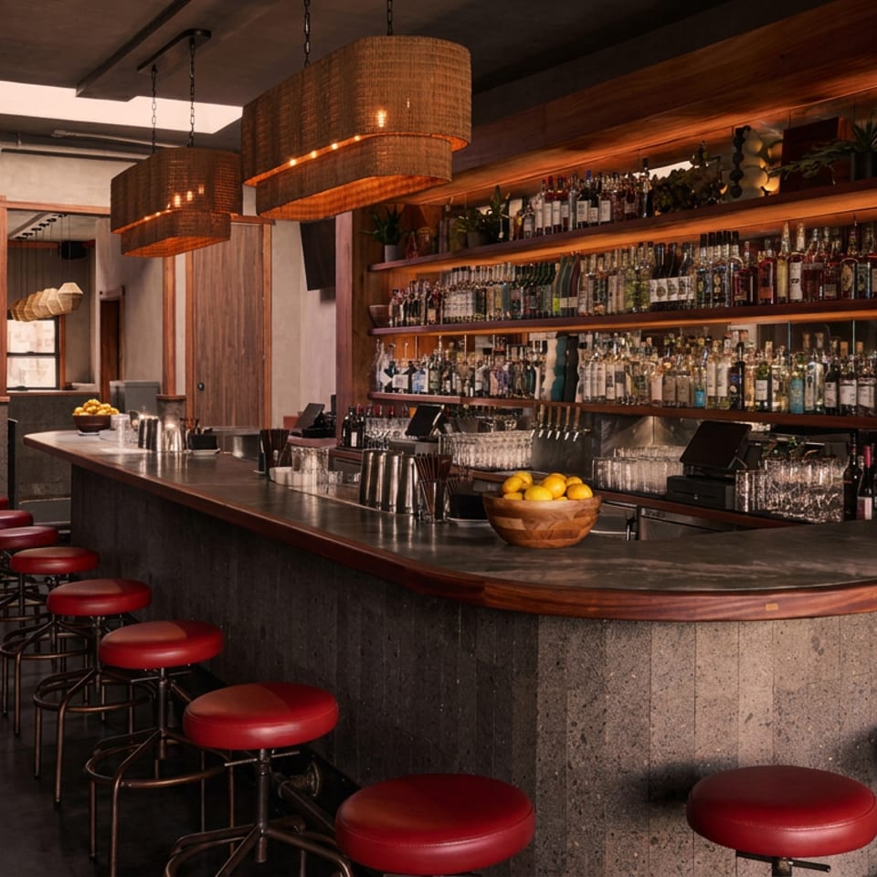 Warmly lit restaurant bar with red stools, wooden shelves of liquor bottles, hanging lamps, and bowls of citrus.