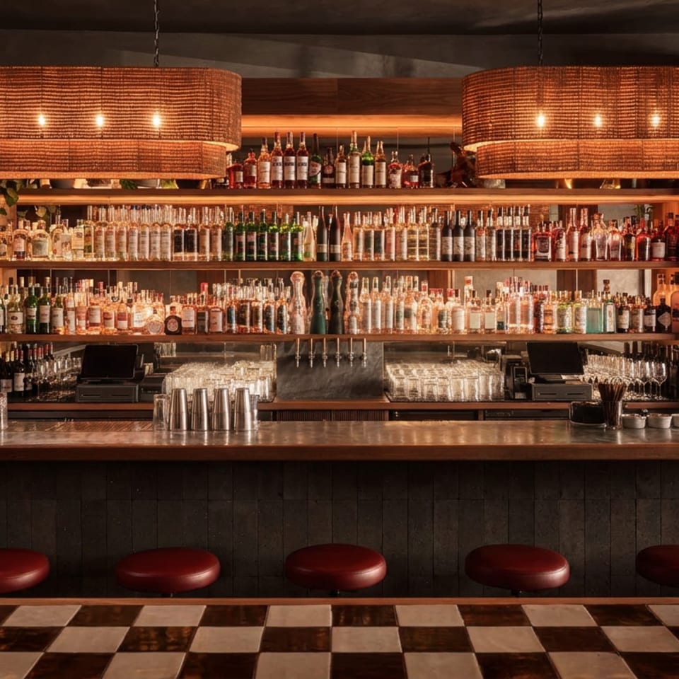 Warmly lit restaurant bar with rows of liquor bottles, hanging woven lamps, copper counter, and red leather stools.