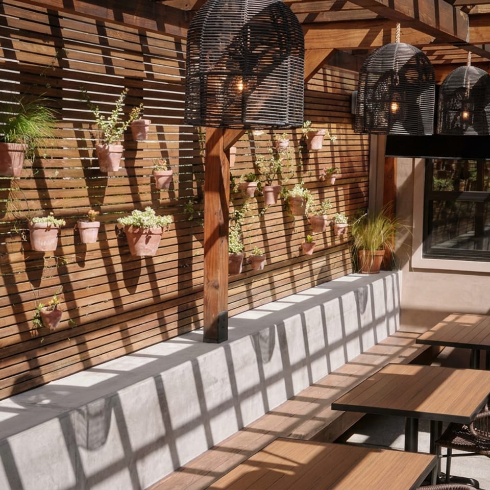 Sunlit restaurant patio with wooden tables, woven chairs, hanging lamps, and a wall decorated with potted plants.