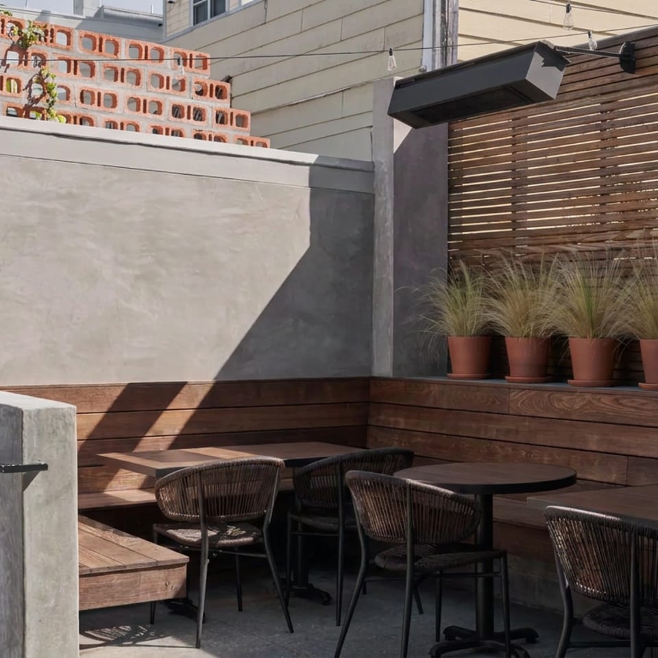 Sunlit restaurant patio with wooden bench seating, woven chairs, tables, potted grasses, and a brick privacy wall.
